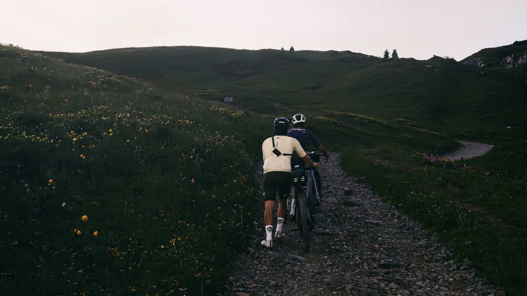 Zwei Radfahrer schieben ihre Gravelbikes mit Bikepacking-Ausrüstung einen felsigen Bergpfad hinauf – aufgenommen in der Dämmerung auf einer abgelegenen Route.
